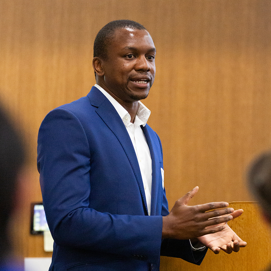 Student in business wear from waist up presenting in front of a wood paneled wall.